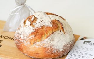 „Frisches Bauernbrot mit Mehl bestäubt auf einem Holzbrett, daneben ein Säckchen Salz und eine Infokarte von RACK IMMO.“